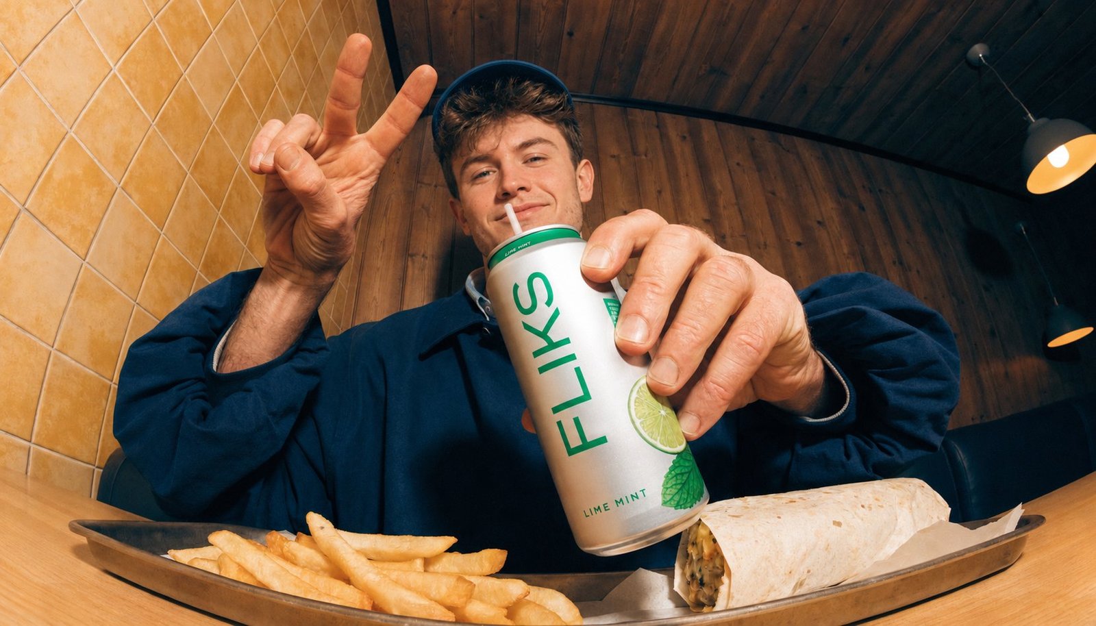Robin fait un signe de la main en tenant une canette Fliks, attablé devant un plateau de frites et un wrap